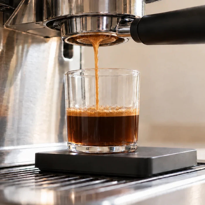 Espresso pouring into a short clear glass containing dark coffee and a thin crema layer.